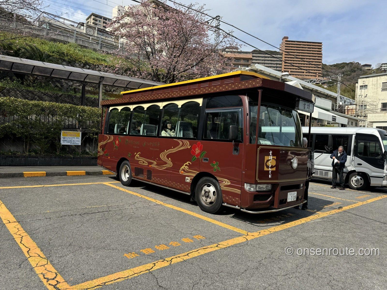 伊豆熱川駅前に停まる吉祥CARENの赤い送迎バス。背景に桜の木