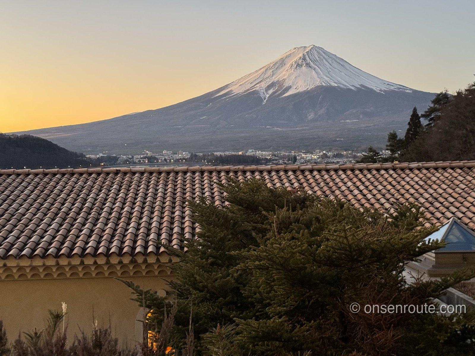 朝焼けの空を背にした雪化粧の富士山。手前にオレンジ色の瓦屋根と針葉樹が見える