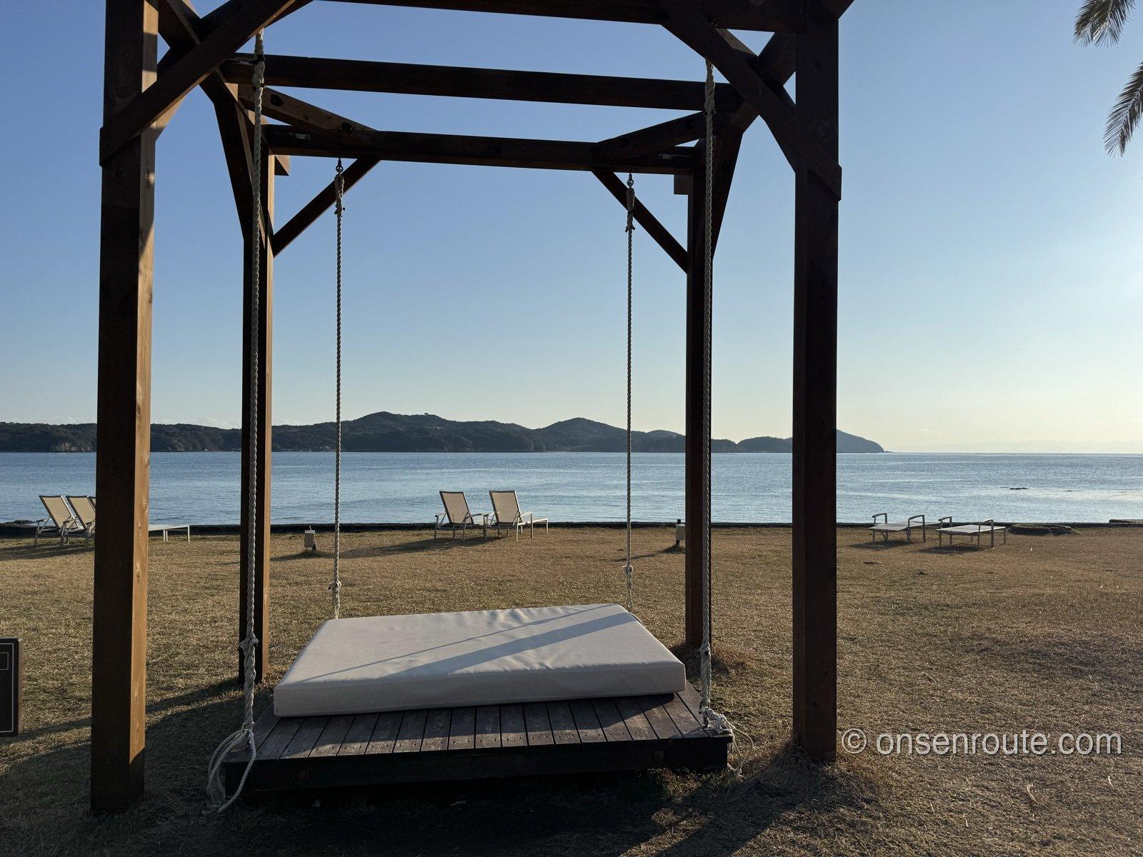 夕日に染まる浜の木組みのデイベッド。ロープで吊られた白いマットの向こうに穏やかな海と対岸の山並み