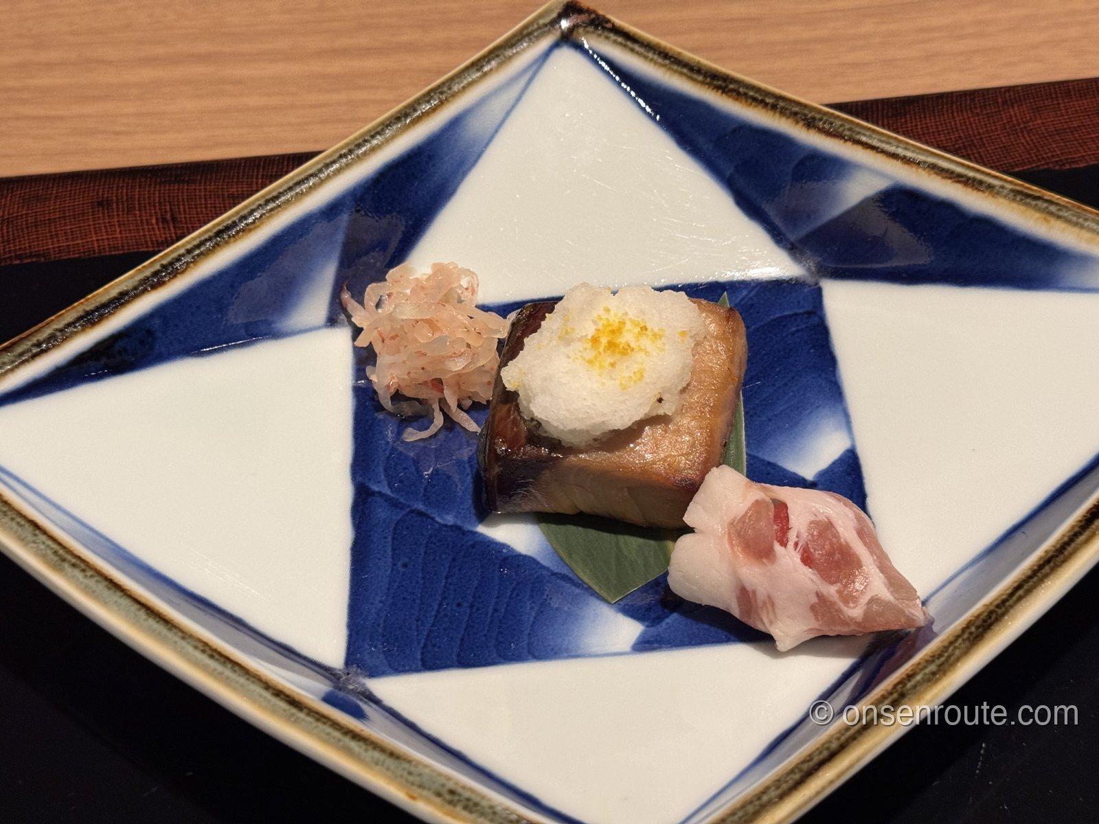 染付の菱形の皿に盛られた焼物。鰤のからすみ焼きの上に白いおろし、左に梅大根、手前に生ハム巻き