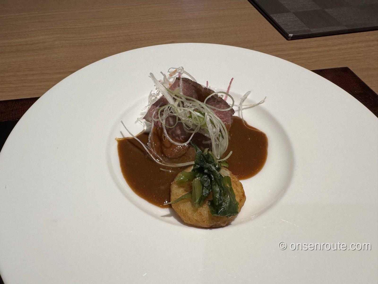 白い皿に盛られた黒毛和牛小春煮。肉の上に白髪葱、じゃが芋餅にわさび菜、味噌デミグラスソース