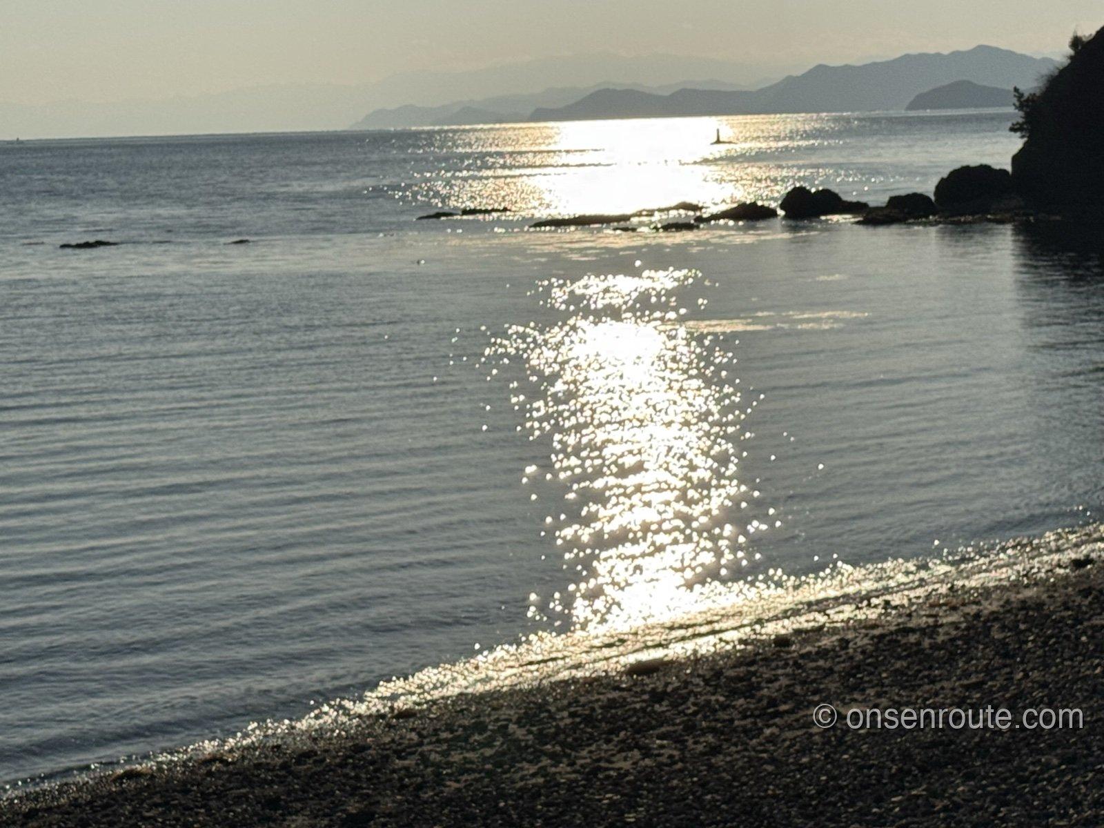 夕方の海面に夕日の光の道がきらめく。手前は玉砂利の浜、奥に岬と山並み