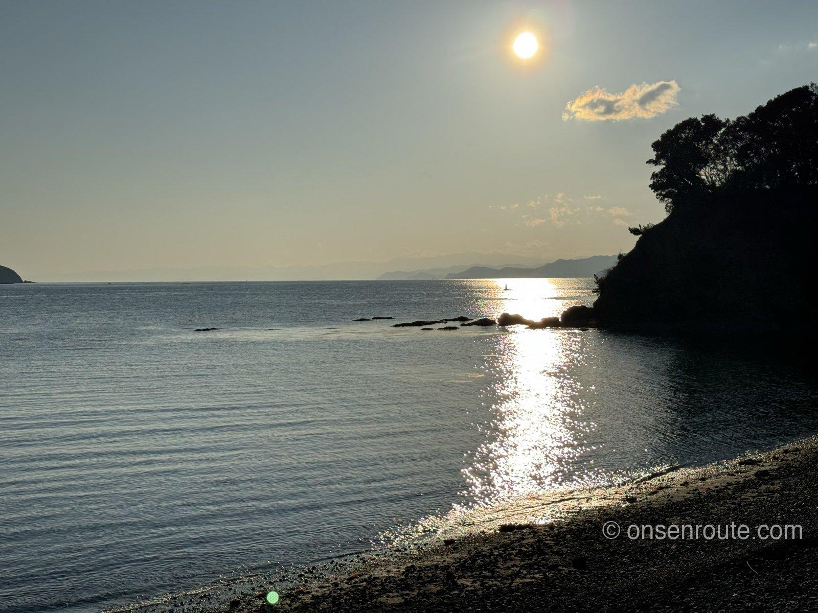 夕日に染まる浜から見る、海に沈みかける太陽。右手に木々の茂る岬のシルエット