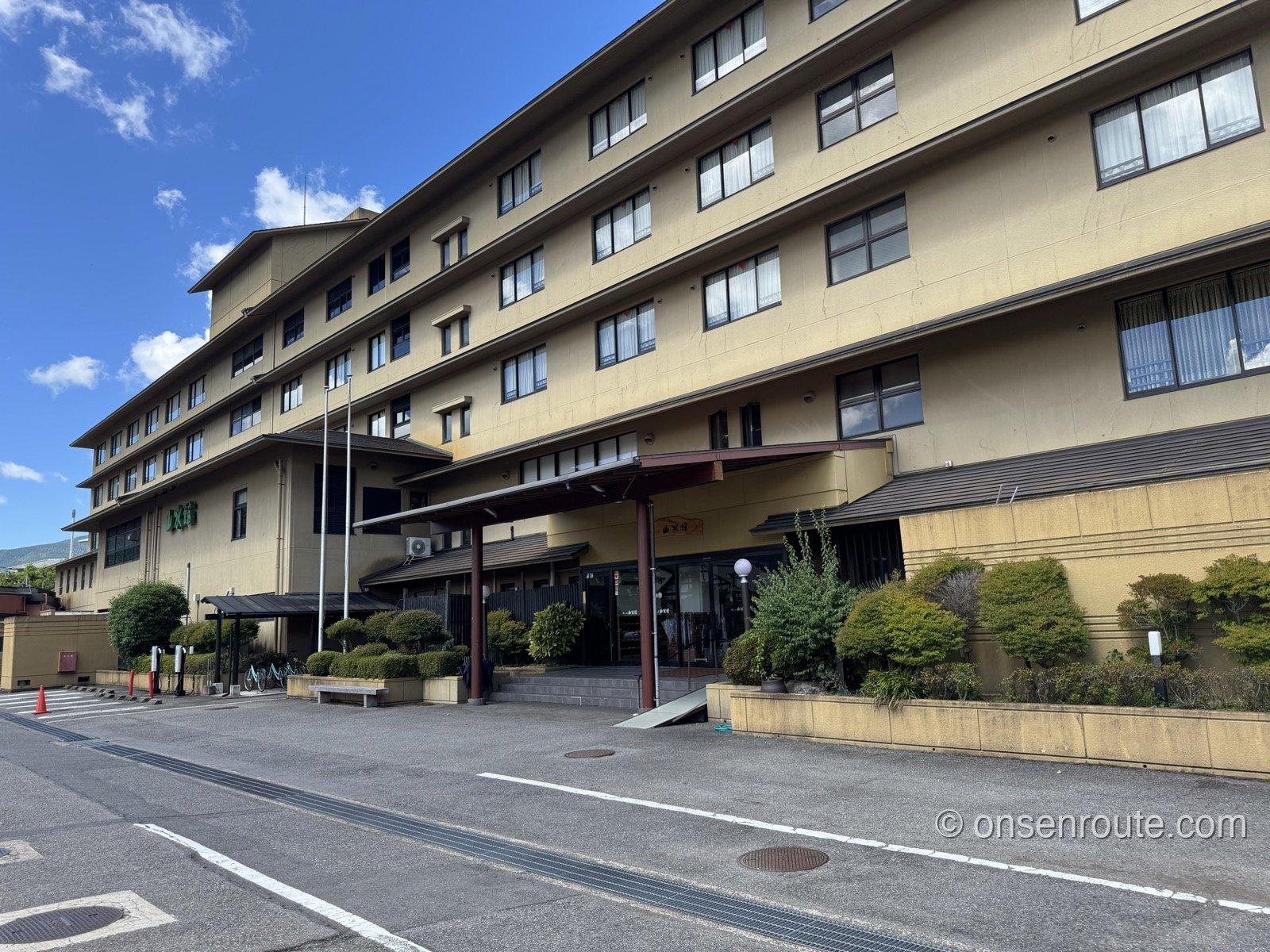 ゆふいん山水館の外観。青空の下、5階建ての大型旅館のエントランスに植栽が並ぶ