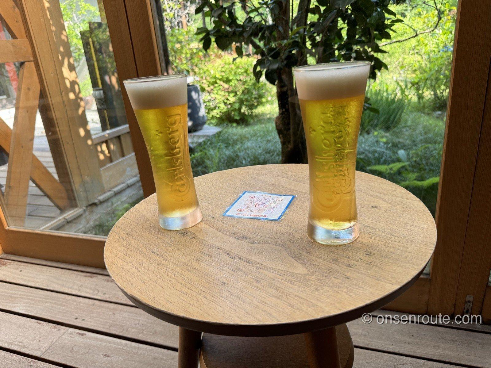 丸いサイドテーブルに置かれた生ビールのグラス2つ。間にコースターが置かれ、窓の外は緑の庭