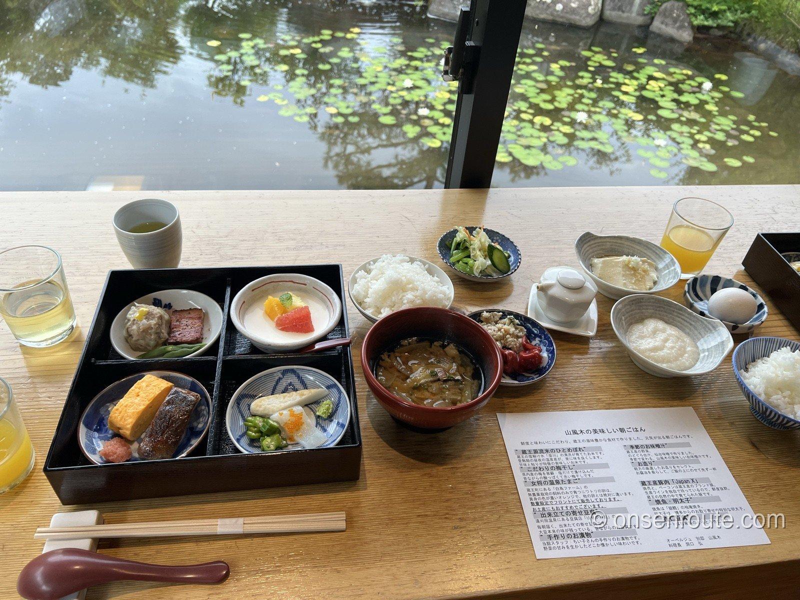 窓際のカウンターに並んだ山風木の朝食。二段の重箱、白飯、味噌汁、寄せ豆腐、とろろ、生卵、お品書きが置かれ、窓の外は睡蓮の浮かぶ池