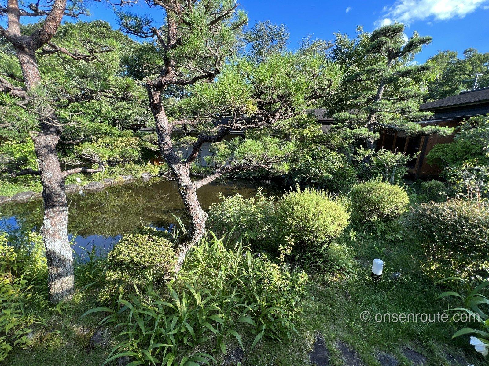 山風木の庭。手入れされた松と刈り込みの植栽に囲まれた池があり、その向こうに黒い屋根の建物が並ぶ。空は快晴