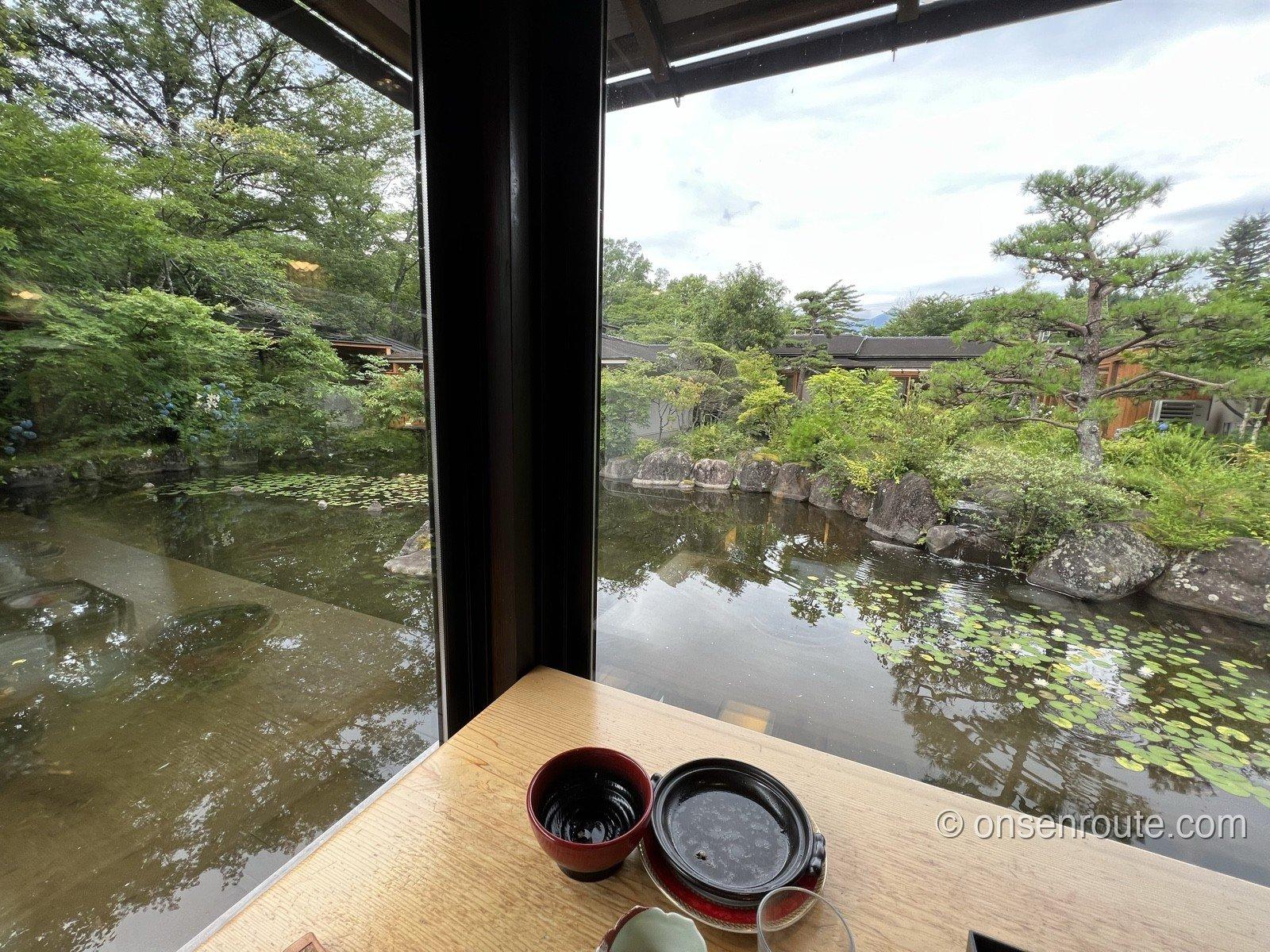 カウンター席の窓から見た池。水面に睡蓮の葉が広がって白い花が咲き、対岸には松と岩、奥に建物の屋根が見える