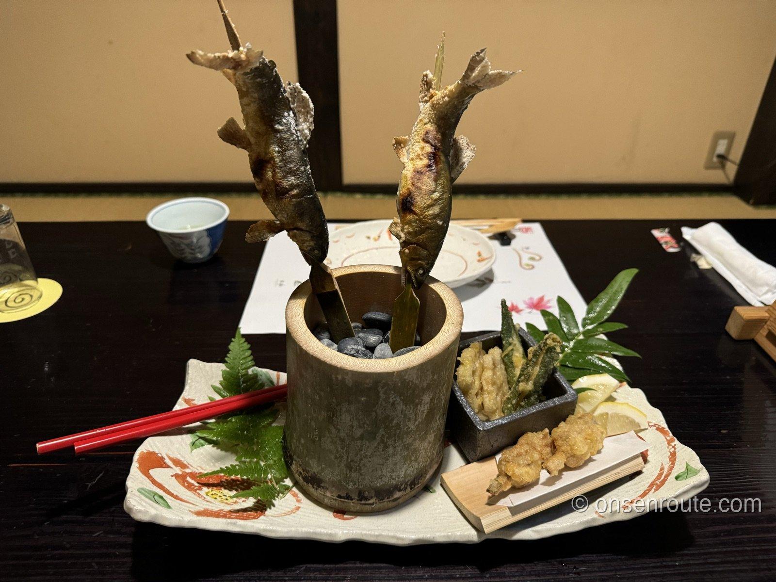 竹筒に立てられた鮎の塩焼き2尾。皿には天ぷらとレモンが添えられている
