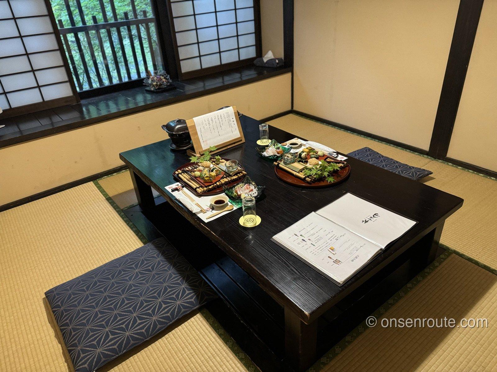 個室の食事処。黒い座卓に前菜の盛り合わせとお品書きが用意され、窓の外に渓流の緑が見える