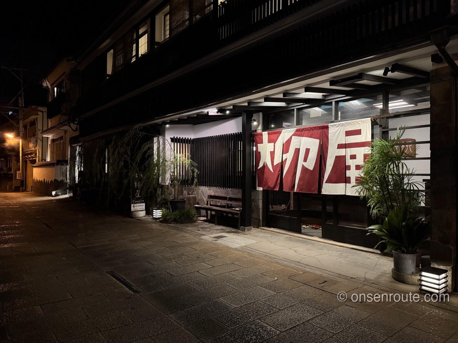 夜の湯治柳屋の外観。暖簾と格子窓に明かりが灯り、石畳の路地が照らされる