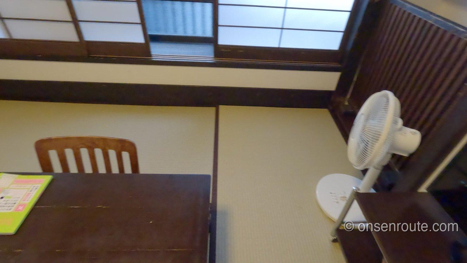 本館湯治部の和室。畳にちゃぶ台と椅子、障子窓、扇風機が置かれたコンパクトな部屋