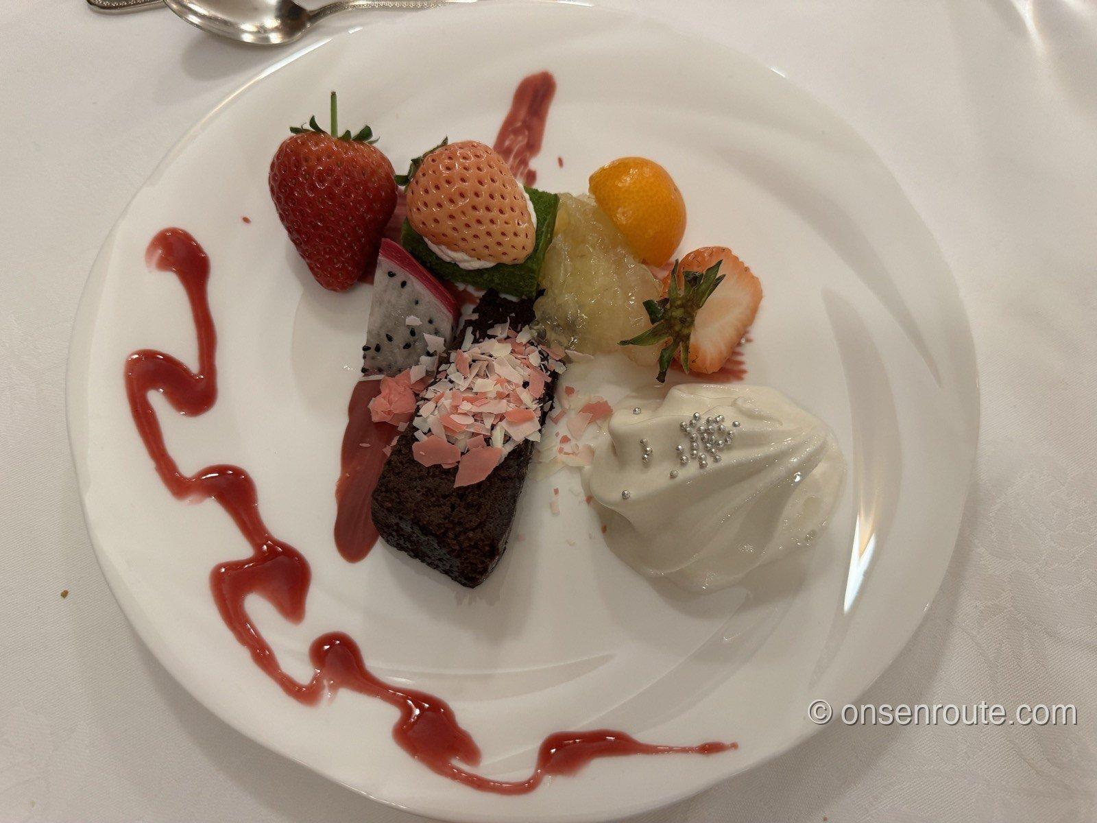 デザートの盛り合わせ。チョコレートケーキに赤と白のいちご、ドラゴンフルーツ、パイナップル、金柑、銀の粒をあしらったクリームが添えられ、皿にベリーソースが曲線で描かれている