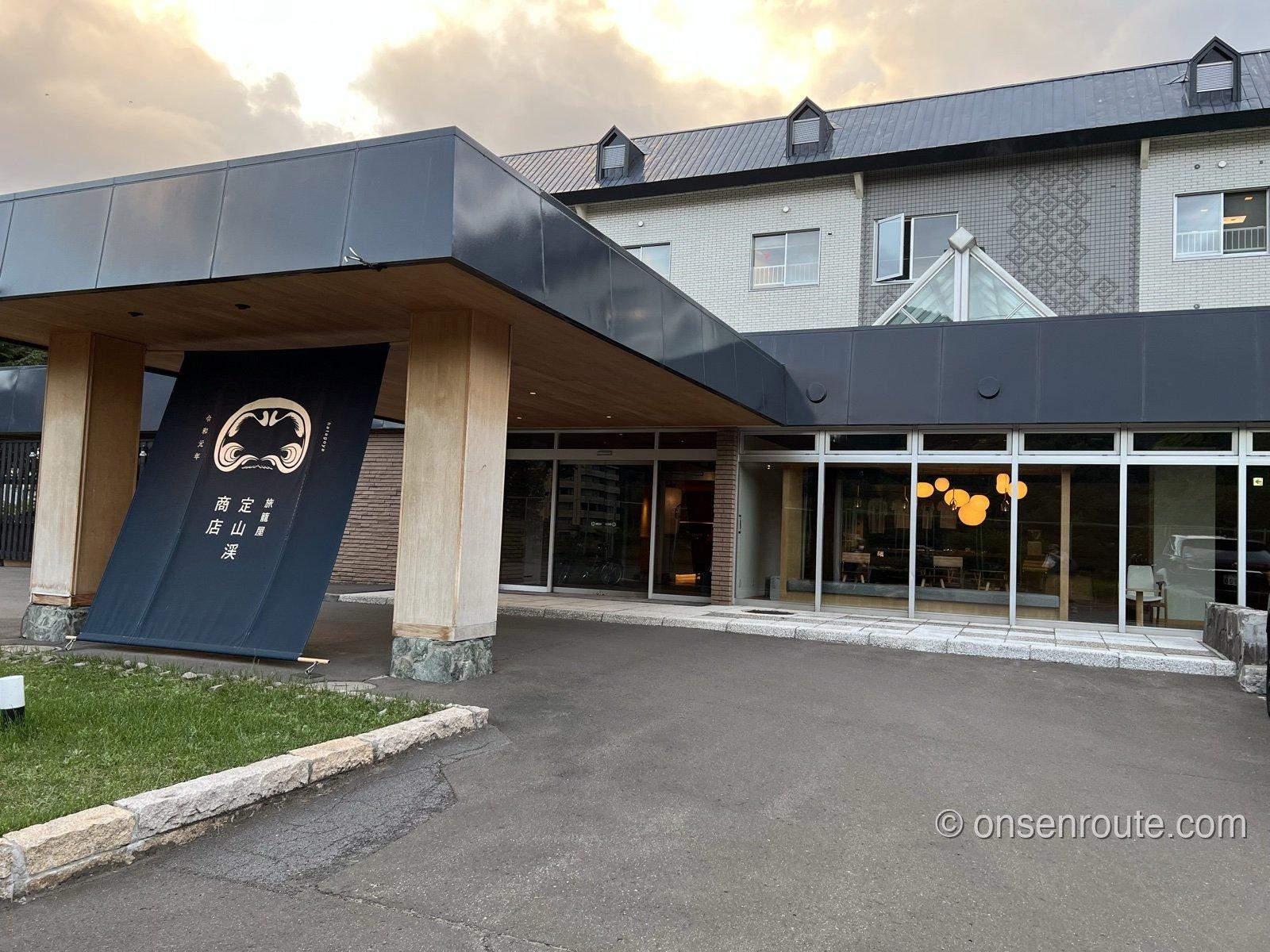 旅籠屋 定山渓商店の外観。夕暮れの空の下、車寄せの脇に宿名と店主の顔を染め抜いた紺色の大暖簾が張られている