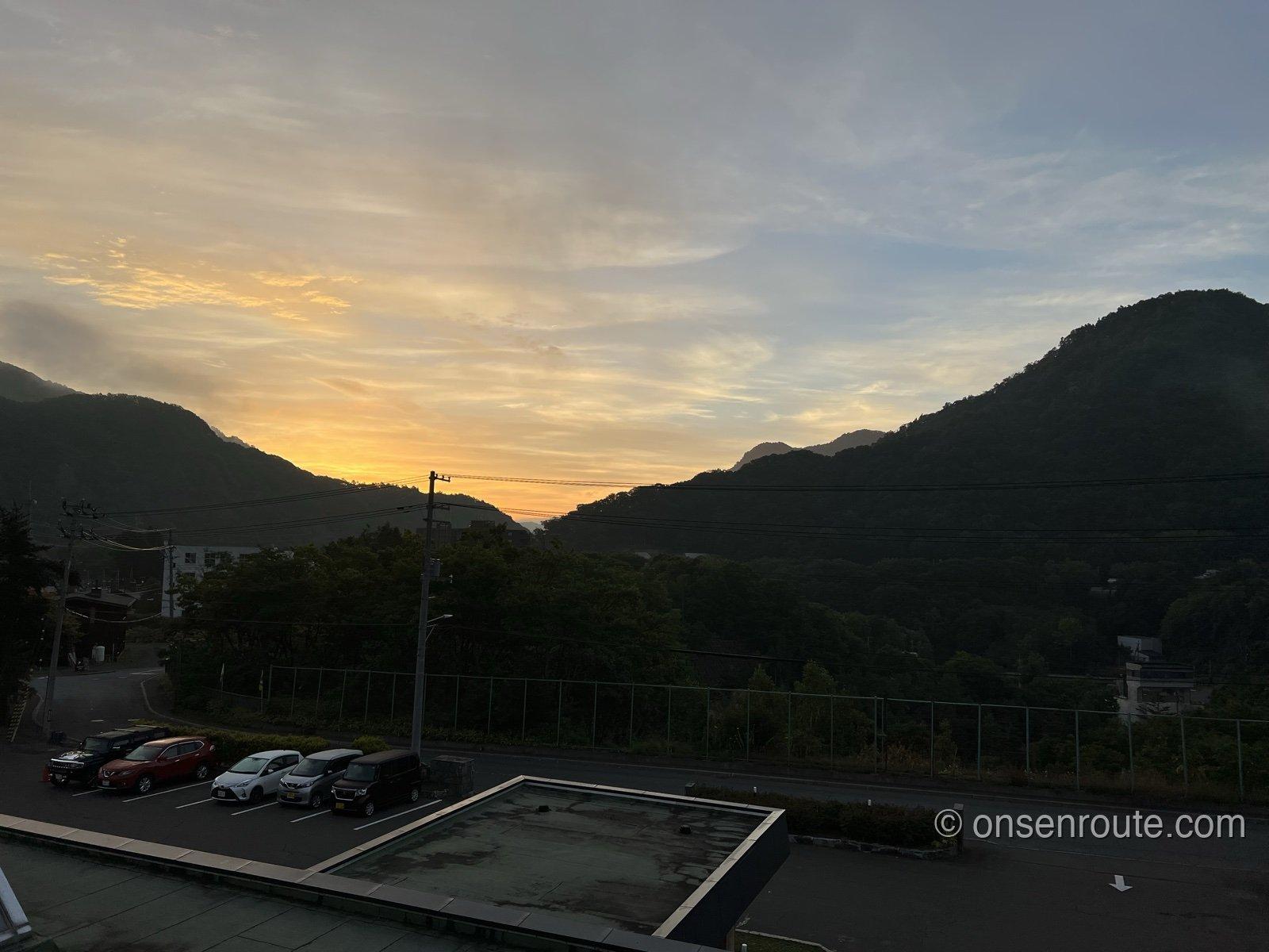 部屋の窓から見た夜明けの定山渓。両側にせまる山の稜線の間の空がオレンジ色に染まり、手前に宿の駐車場が見える
