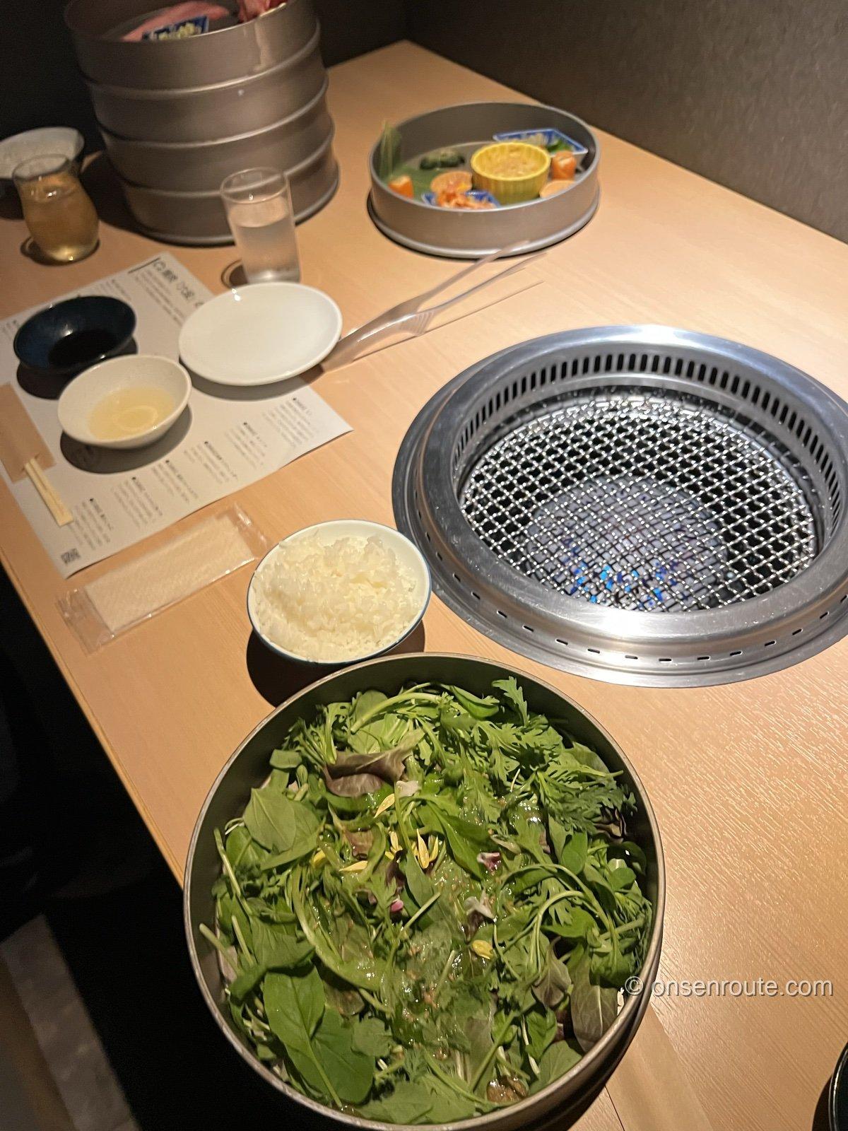 焼肉のテーブル。中央に七輪、手前に山盛りの葉物サラダとご飯、奥に前菜のトレーとタレの小鉢が並ぶ