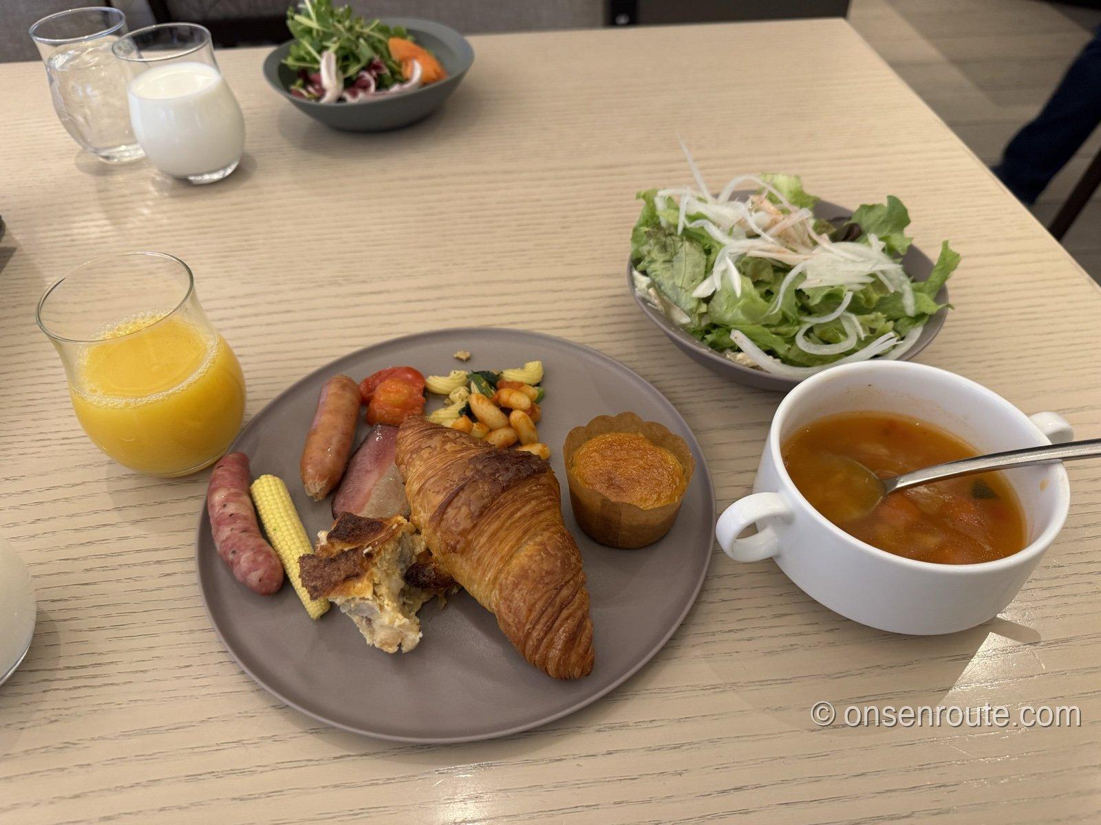 朝食ビュッフェの盛り付け。クロワッサン、ソーセージ、ヤングコーン、スープ、サラダ、オレンジジュース