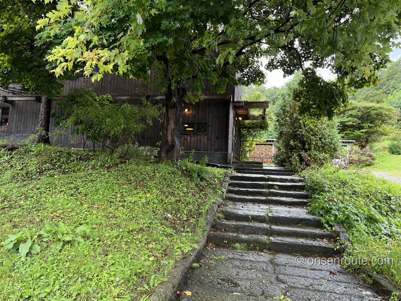 緑の丘に建つエグ・ヴィヴの黒い木造店舗。大木の脇から石段が入口へ続く