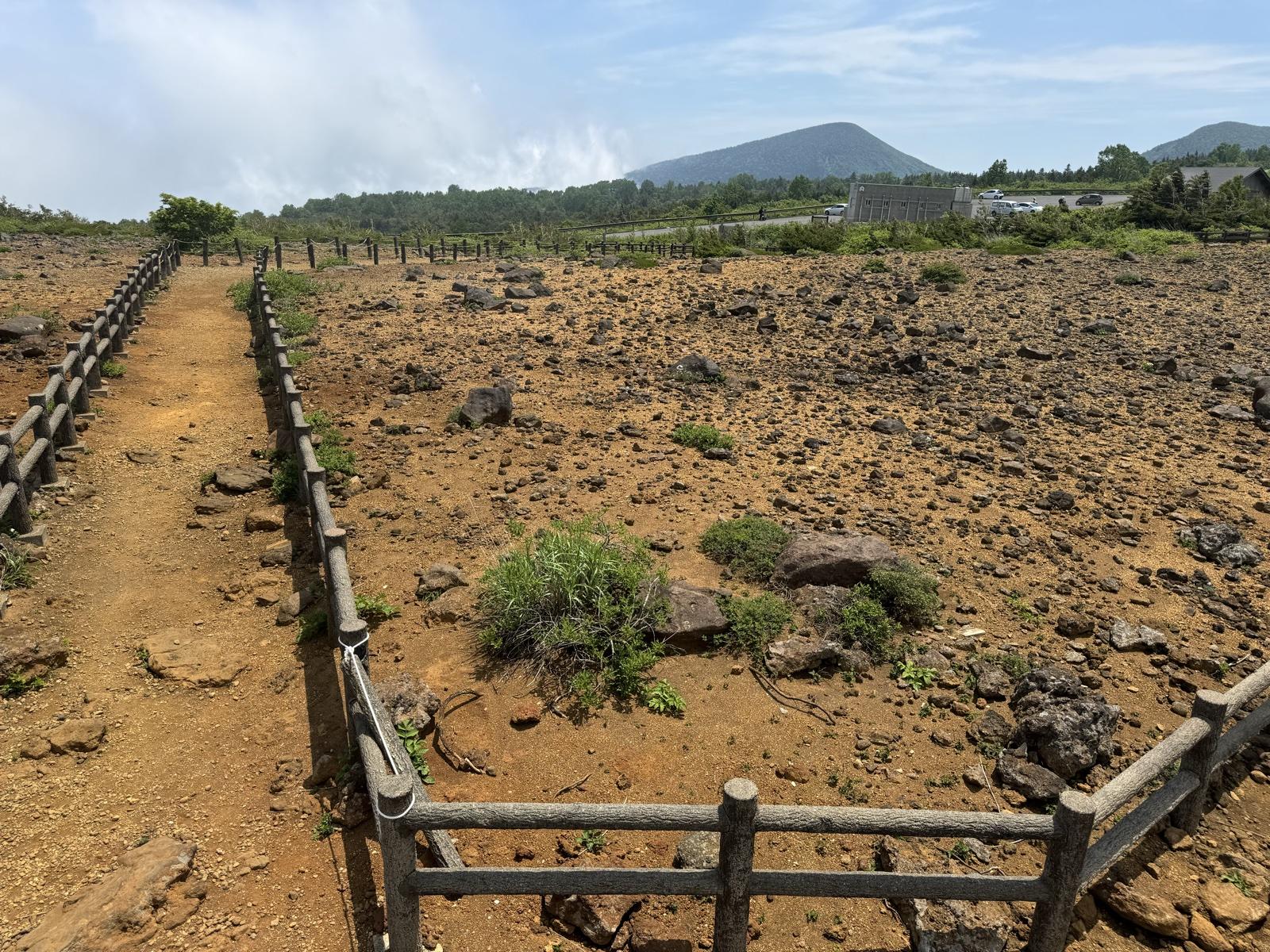駒草平の火山礫の荒野。柵に沿って遊歩道が延び、遠くに蔵王の山並みと駐車場が見える
