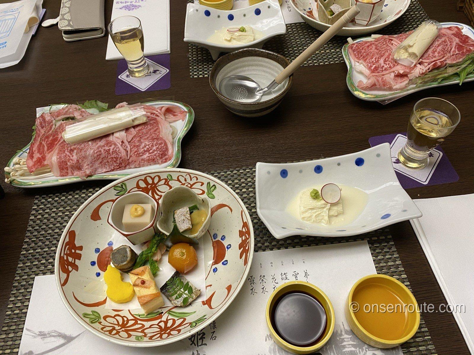 懐石の夕食。霜降り和牛の薄切りを盛った皿と鍋、彩り豊かな前菜の盛り合わせ、湯葉の小鉢、梅酒のグラスが並ぶ