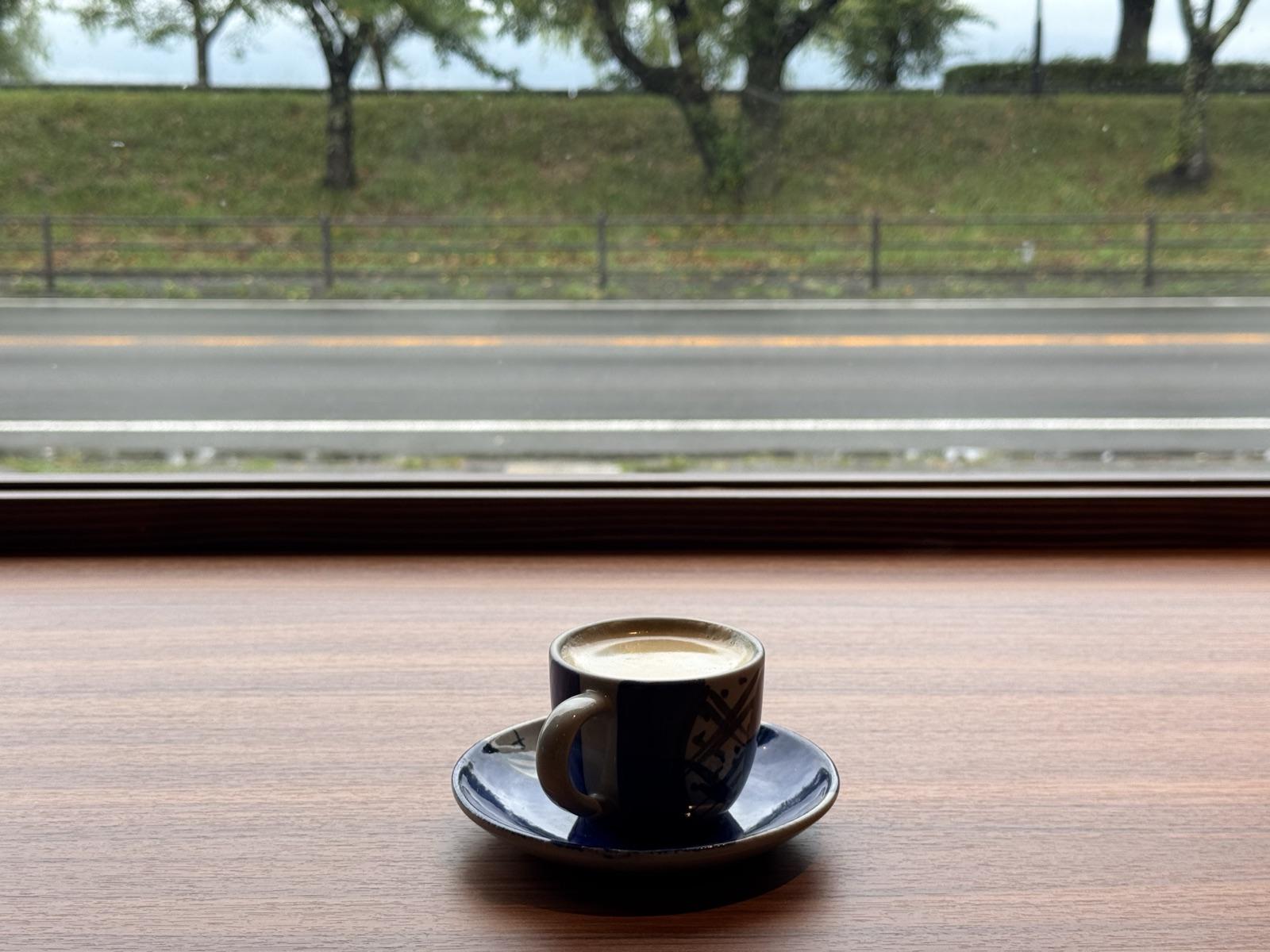 窓際のカウンターに置かれた藍色のカップのコーヒー。窓の外に湖畔の道路と並木
