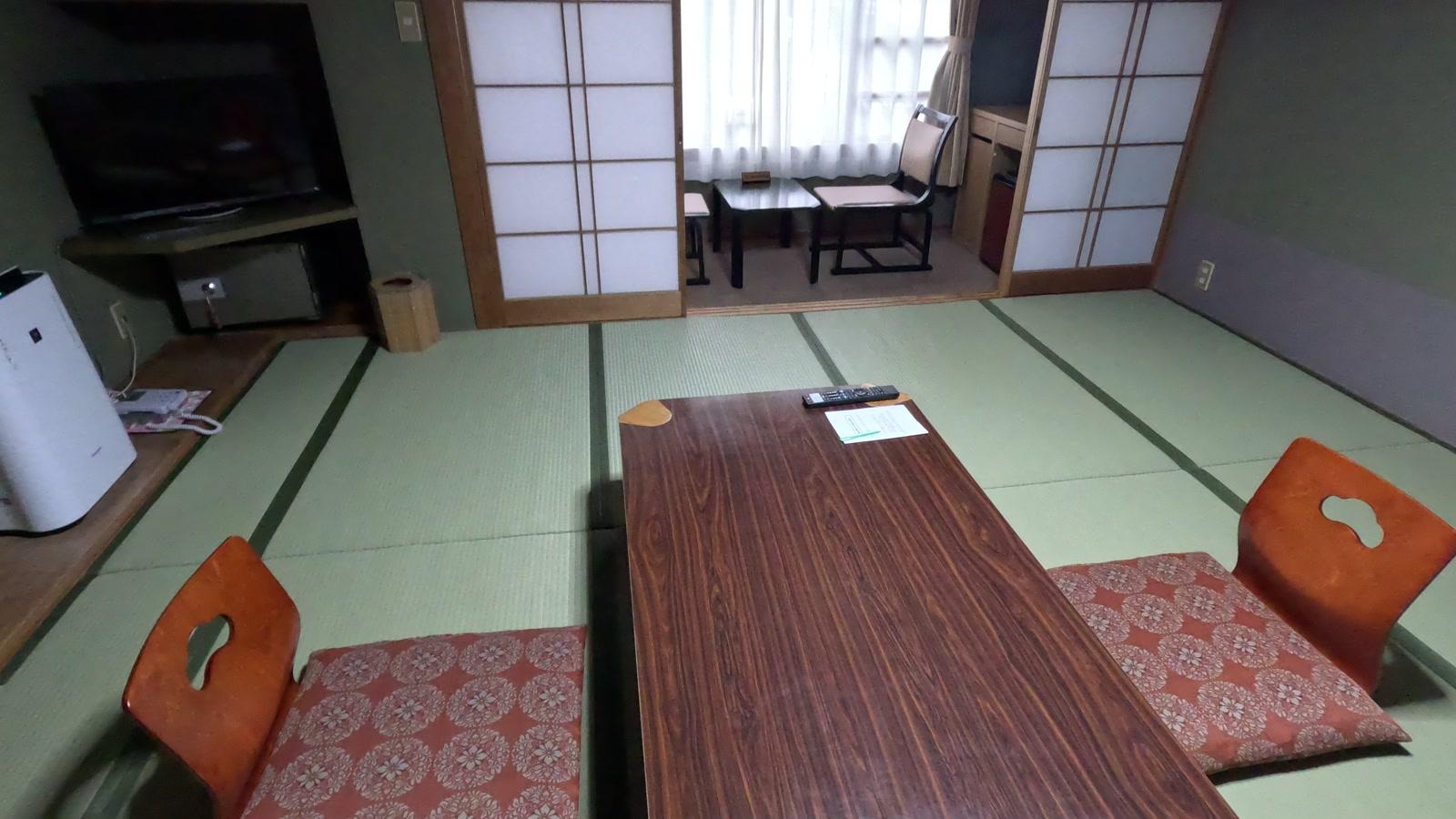 すわ湖苑の和室12畳。座卓と座椅子、左にテレビと空気清浄機、奥の障子の向こうに広縁のテーブルと椅子