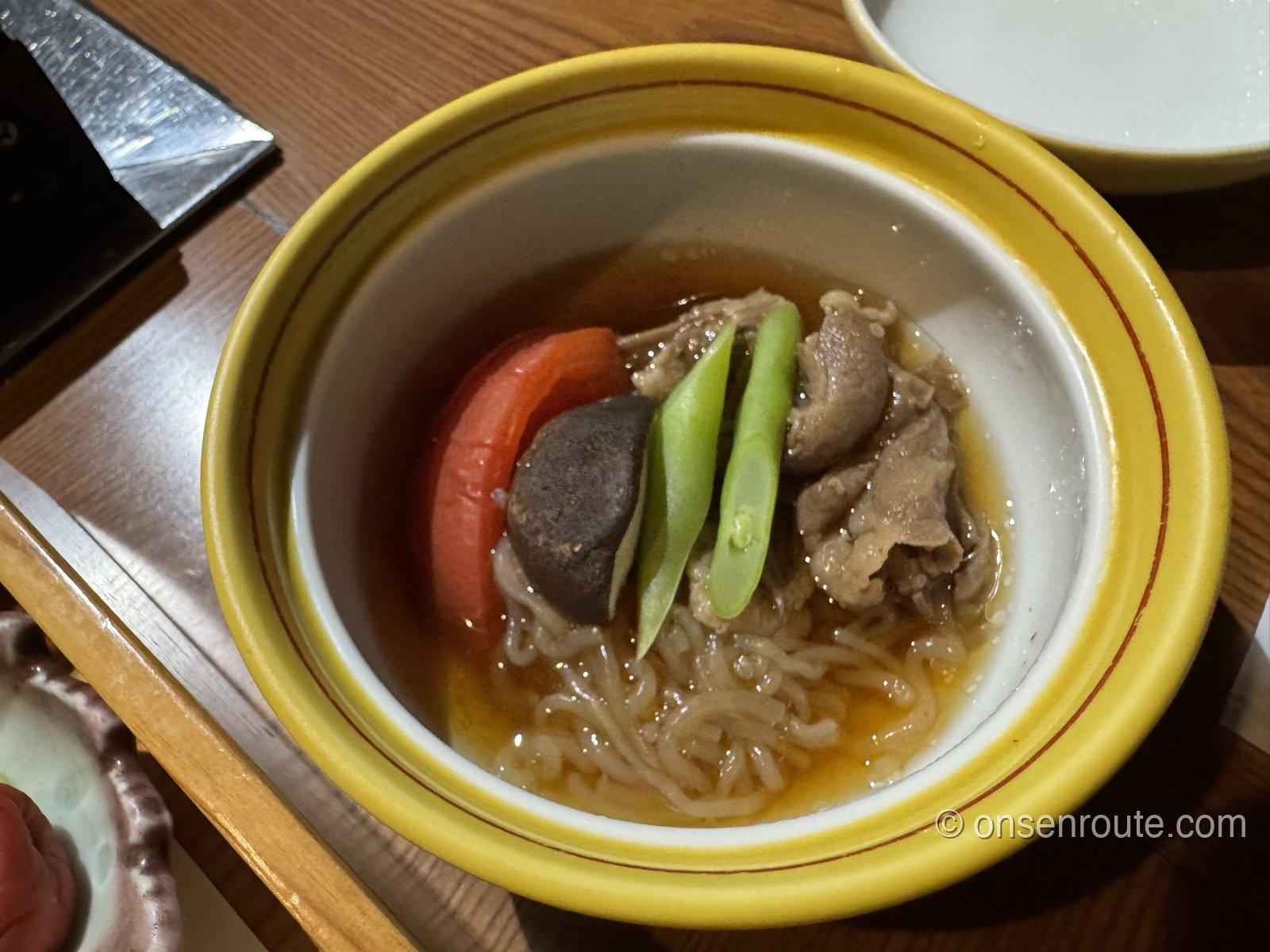黄色い縁の椀に入った芋煮風の温かい一品。牛肉、しいたけ、いんげん、トマト、しらたきが出汁に浸かる