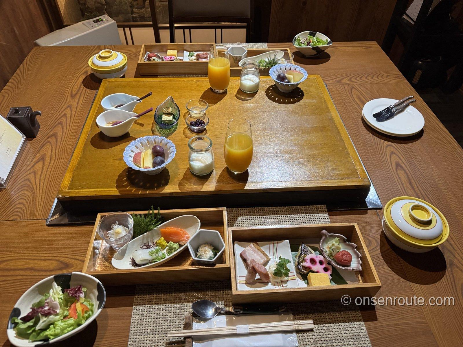 朝食の卓。木の箱膳に刺身や卵焼き、焼き魚、梅干しなどの小鉢が並び、ジュースやヨーグルト、フルーツも揃う