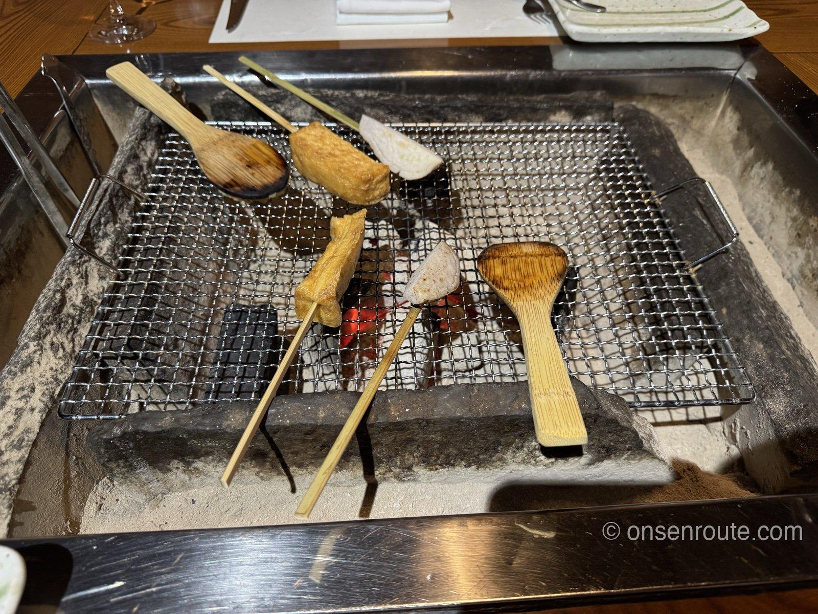 卓上の囲炉裏。網の上に油揚げやえびいもの串が並び、炭火が赤く熾きている