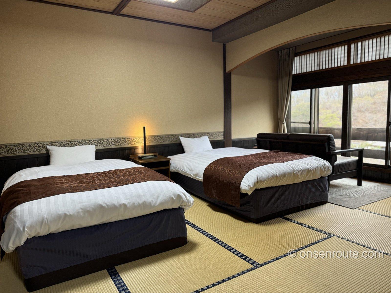 安達屋の客室。畳敷きの部屋にツインベッドが並び、窓際にソファ。壁は生成りの和紙調で腰壁は焦げ茶