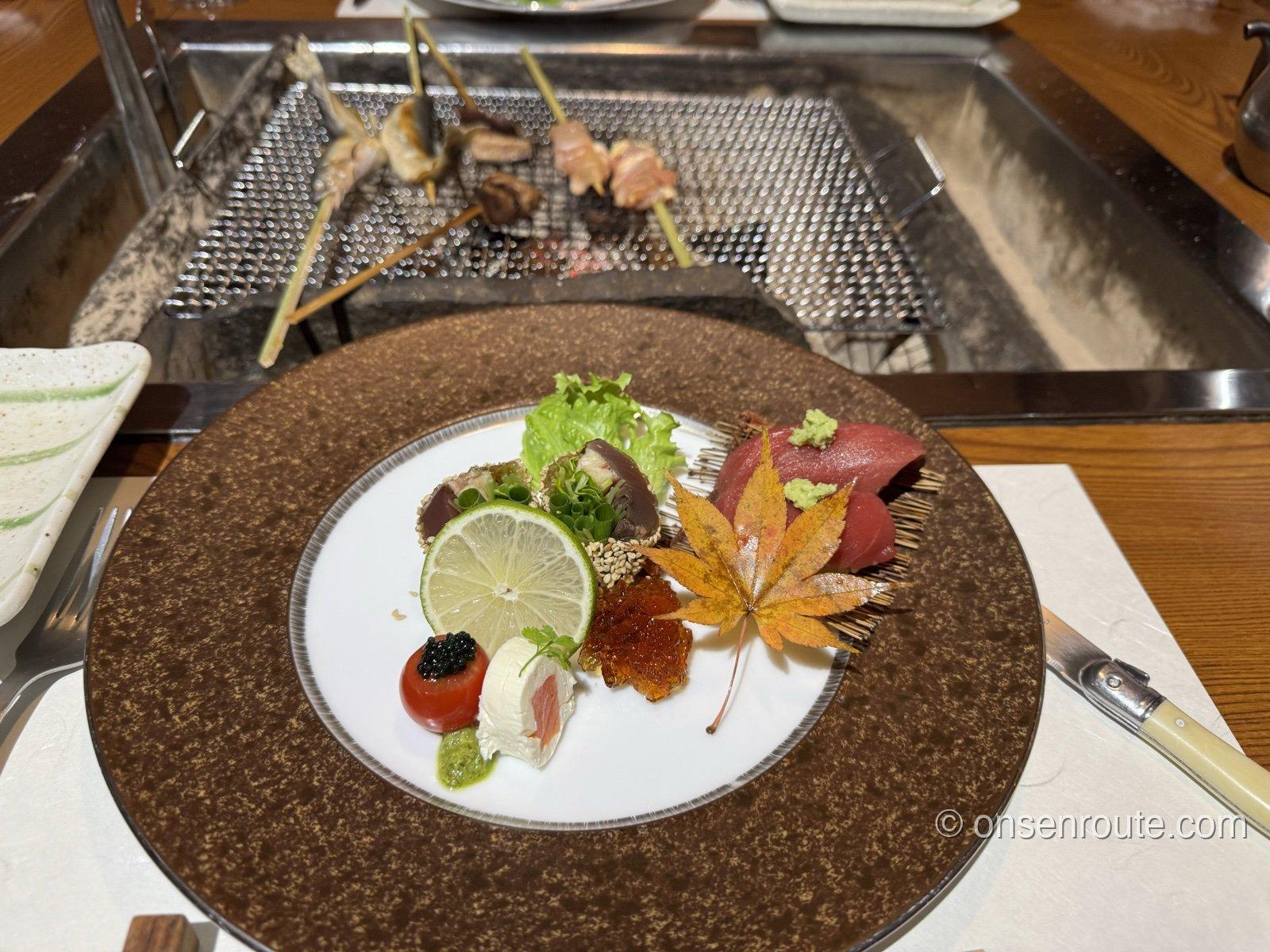 夕食のお造り。茶色の大皿に、まぐろ、炙りのかつお、いくら、トマトとチーズの一品が紅葉の葉とともに盛られる。奥の囲炉裏ではニシンや鮎、鶏の串が焼ける