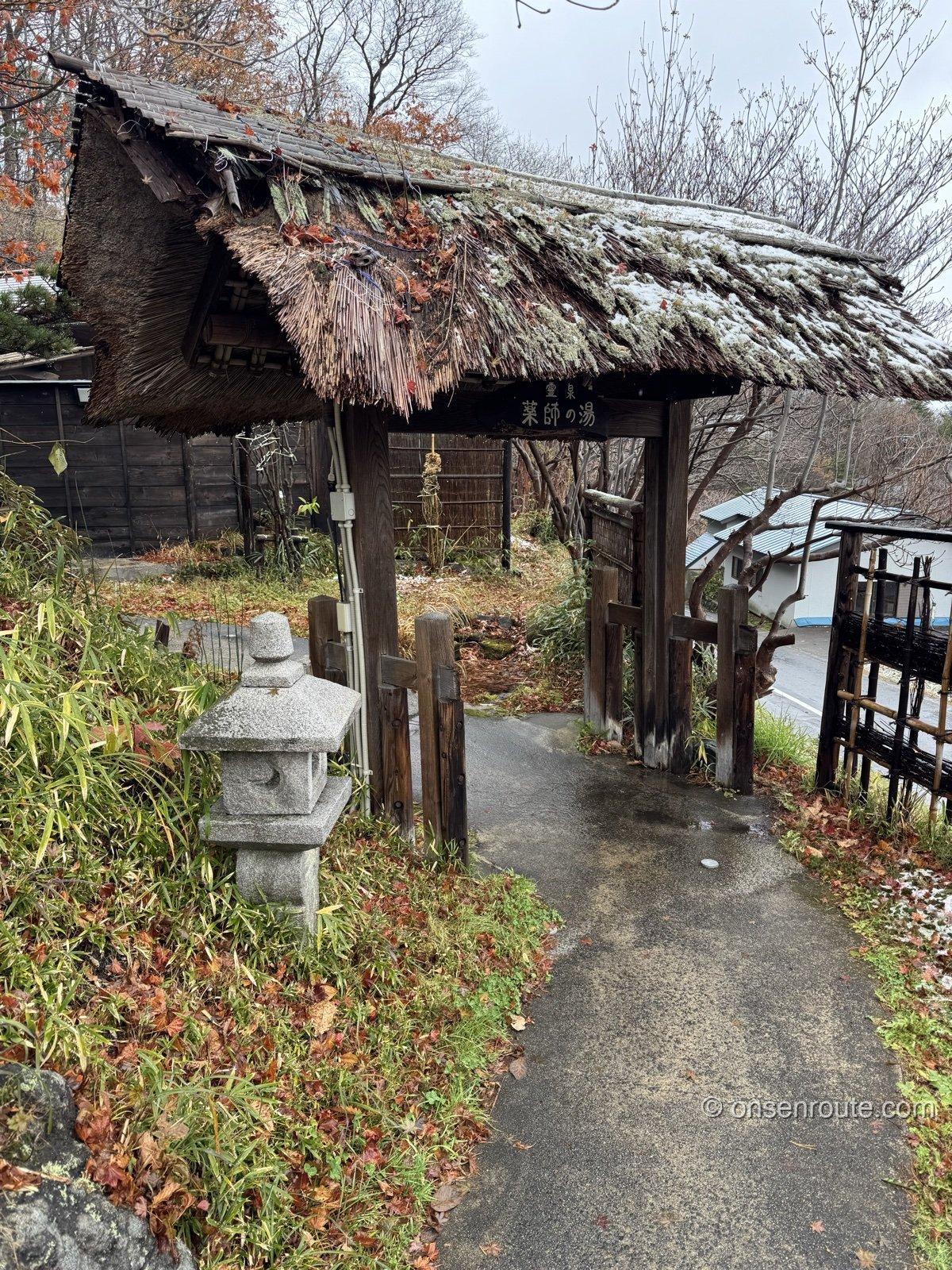 「霊泉 薬師の湯」の看板を掲げた茅葺き屋根の門。石灯籠と落ち葉の小道が続く