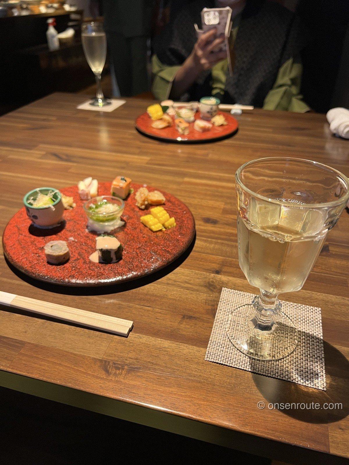 大忠の夕食の前菜。赤い丸皿に小鉢がいくつも並び、手前にとうもろこし、奥にスパークリングワインと白ワインのグラスが置かれている