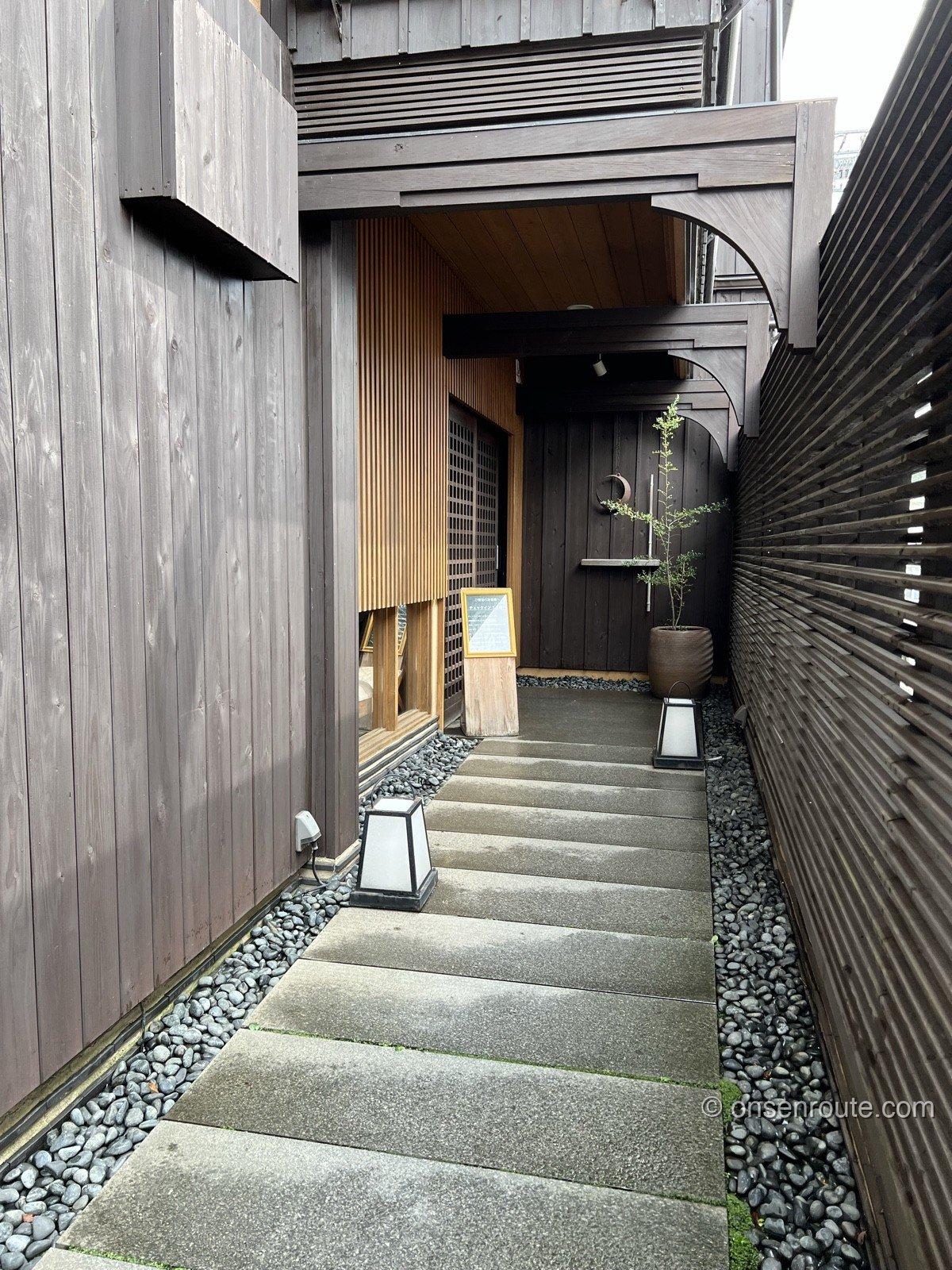 大忠の玄関へのアプローチ。木の梁を渡した屋根の下に石畳が伸び、両脇に玉砂利と石積み、奥に格子戸が見える