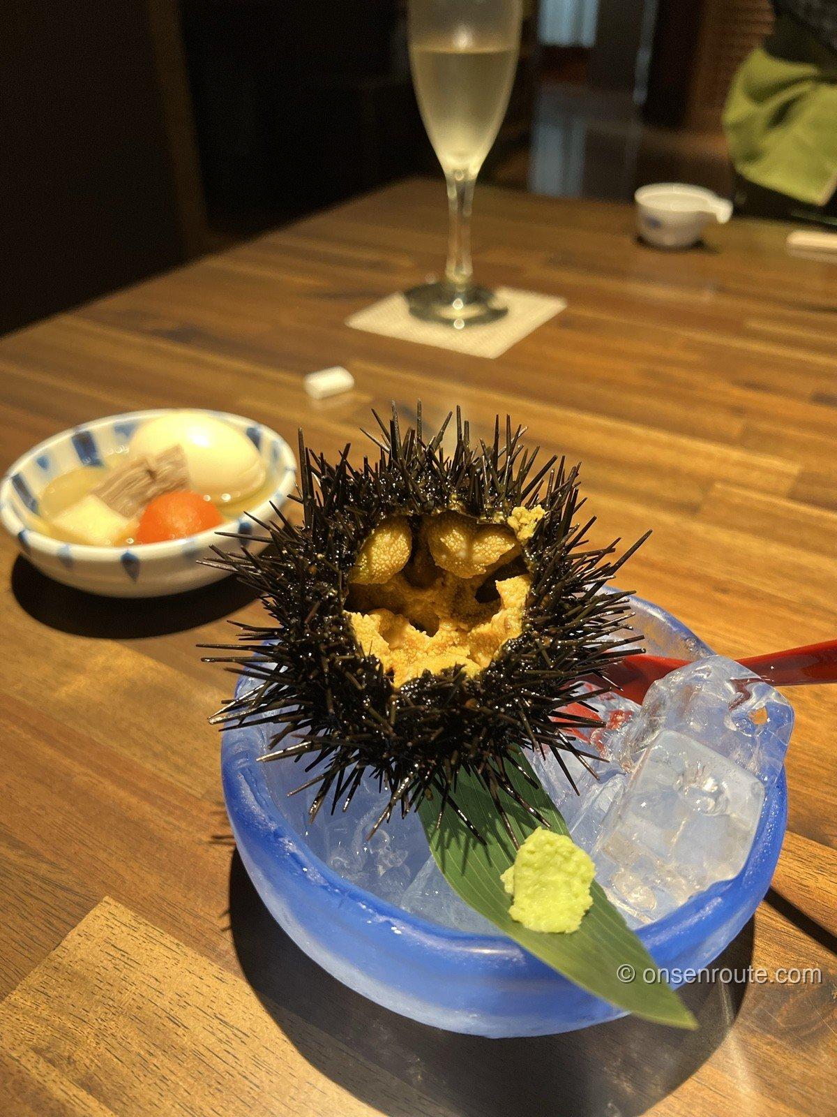 殻付きの生うに。とげのある黒い殻の中にオレンジ色の身が並び、手前におでんの小鉢とスパークリングワインのグラスが置かれている