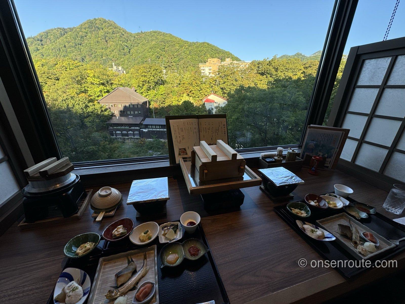 大きな窓の外に緑の山と温泉街の建物が見える席に、朝食の膳が並ぶ
