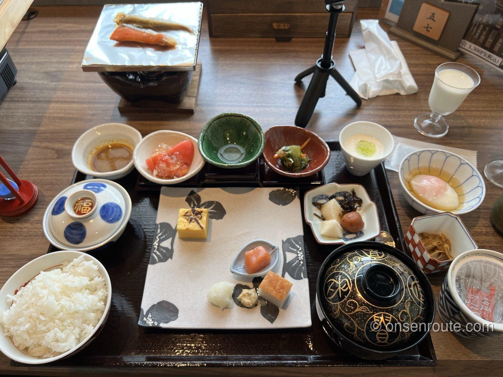 朝食の膳。ご飯と蓋つきの椀、小鉢が数多く並び、奥の焼き台で鮭とししゃもを炙る