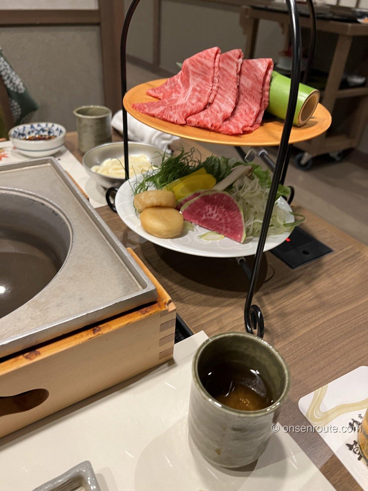 夕食の卓。二段の台に牛肉の皿と野菜が載り、手前に石の鍋が湯気を立てる