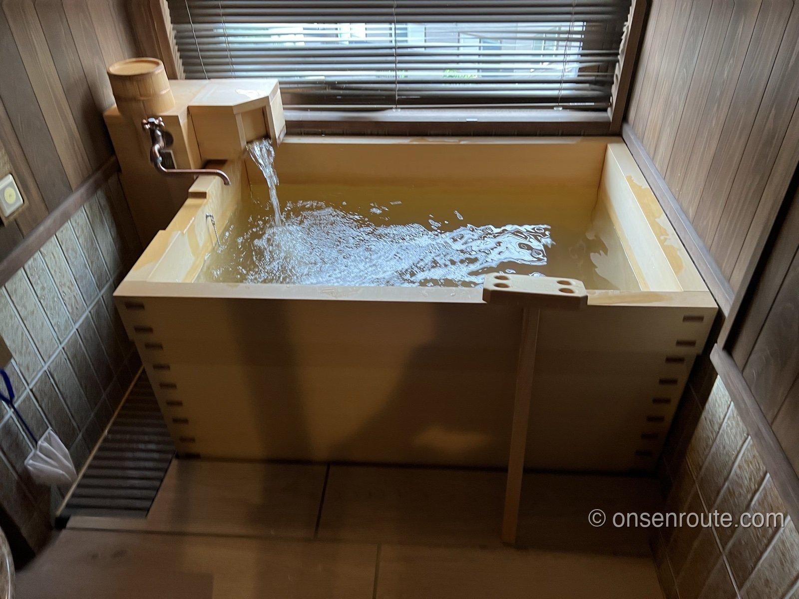 客室の温泉風呂。木の湯船に湯口から勢いよく温泉が注がれている。窓の外にブラインド越しの光