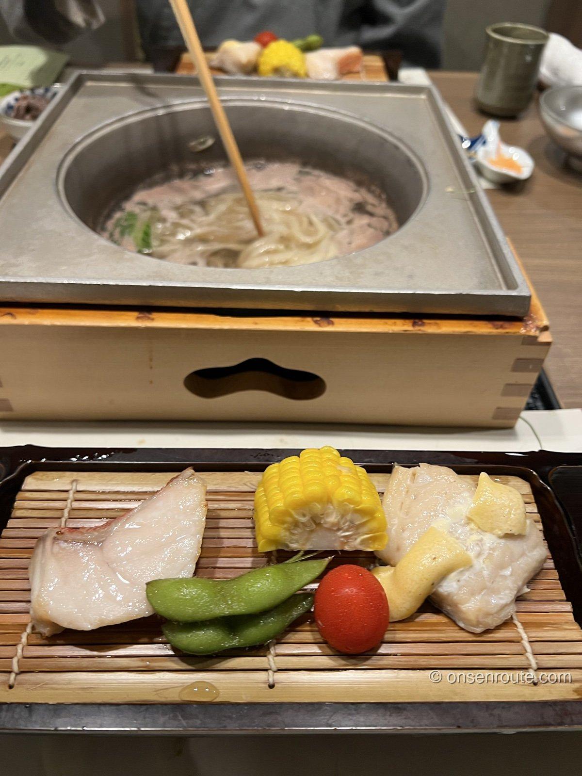 石の鍋でしゃぶしゃぶをする様子。だしの中に肉と野菜、うどん。手前の簀の子に焼き魚ととうもろこし、枝豆