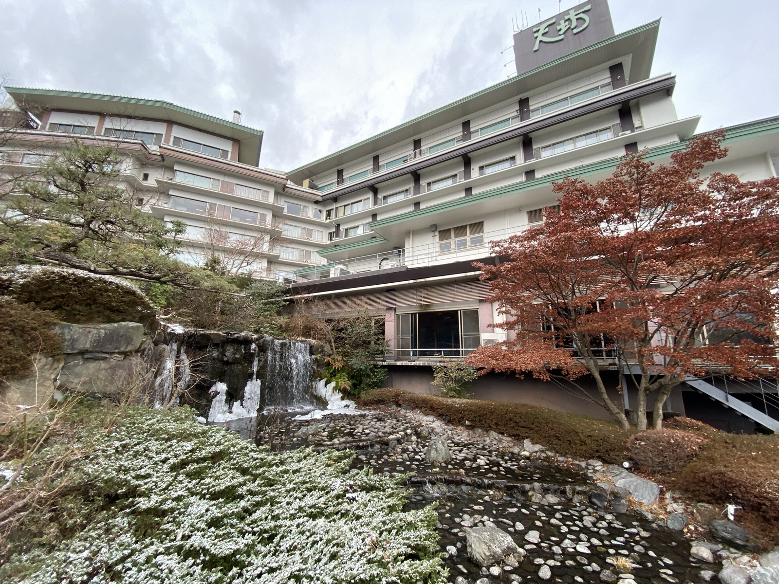 伊香保温泉 ホテル天坊の外観。庭園の滝と雪の残る植え込みの向こうに大型の宿泊棟が建つ