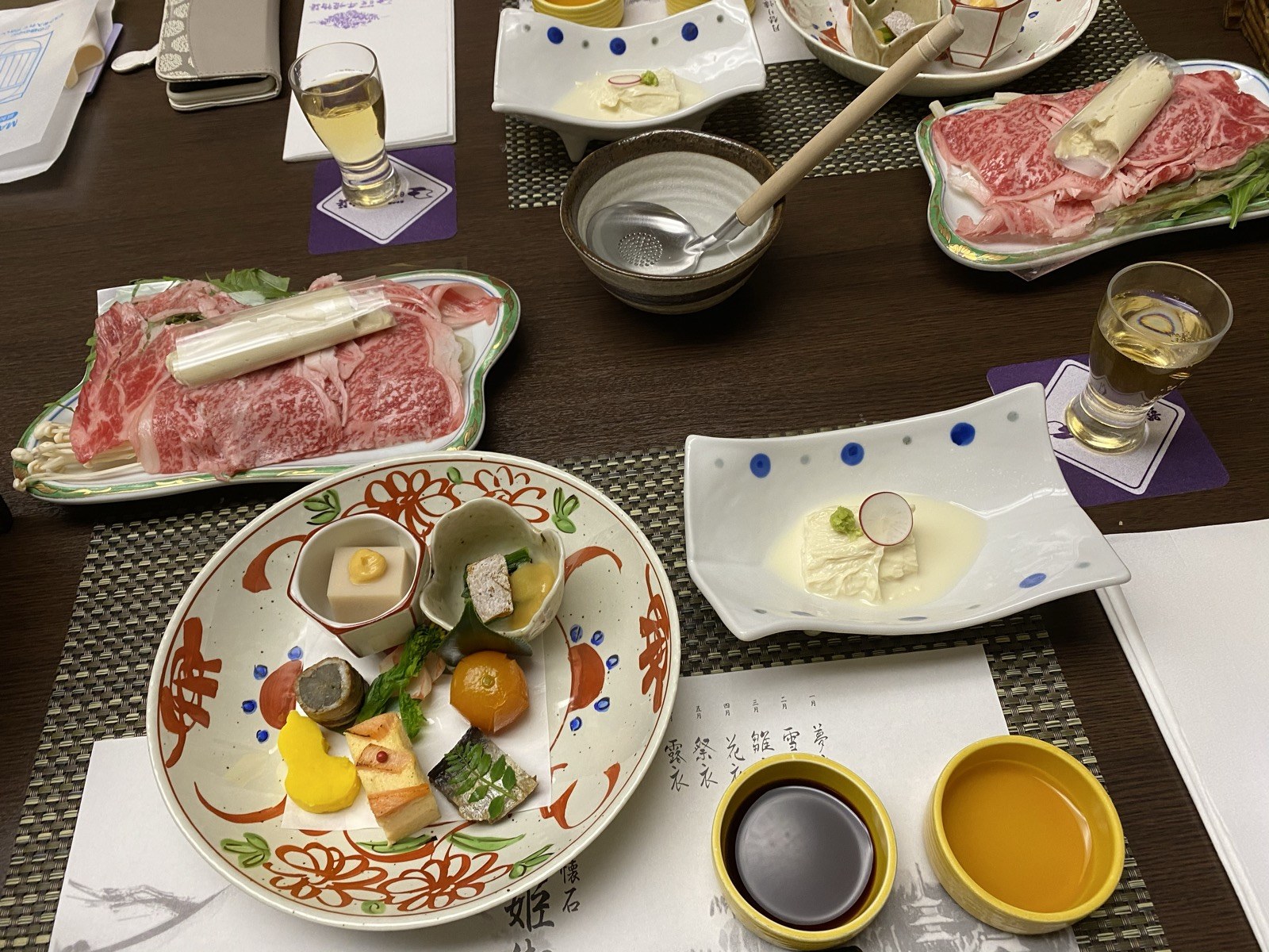 懐石の夕食。霜降り和牛の薄切りを盛った皿と鍋、彩り豊かな前菜の盛り合わせ、湯葉の小鉢、梅酒のグラスが並ぶ